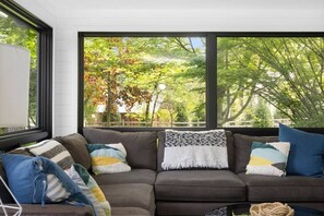 A modern living room with a sectional sofa, large windows, and a view of trees. - River's Edge (Leland)