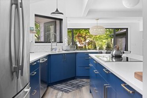 A modern kitchen with blue cabinets, a stainless steel refrigerator, and a large window overlooking greenery.