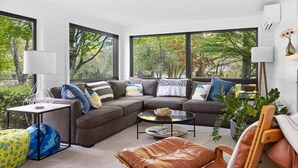 A modern living room with a sectional sofa, a coffee table, and a view of trees through large windows. - River's Edge (Leland)