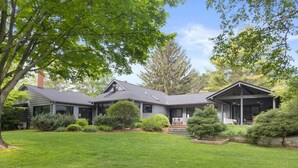 A modern house with a well-maintained lawn, a covered porch, and a large tree in the foreground. - River's Edge (Leland)
