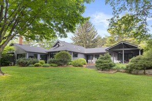 A modern house with a well-maintained lawn, a covered porch, and a large tree in the foreground. - River's Edge (Leland)