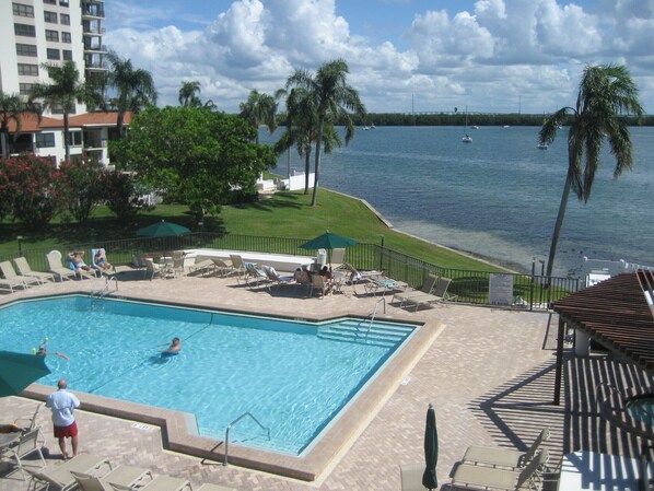 A pool area with lounge chairs and umbrellas, a view of the water, and a building in the background.