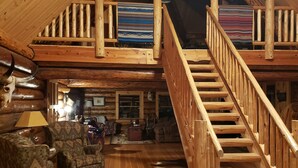 Interior espacioso de una cabaña de troncos con una escalera de madera que conduce a una zona de entrepiso, una chimenea de piedra y un rincón de asientos acogedor.