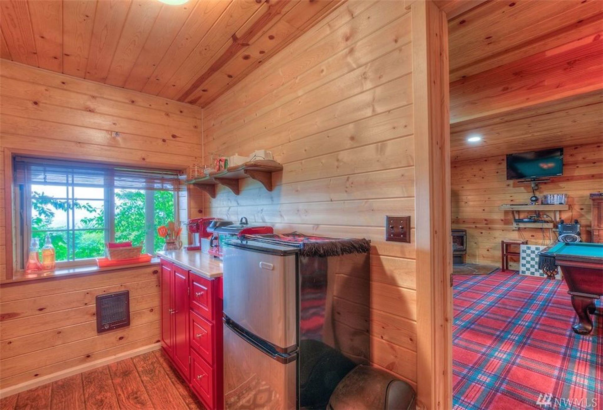A cabin interior with a kitchen area featuring red cabinets, a stainless steel refrigerator, and a window with a view of greenery.