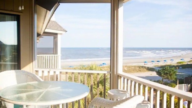 Ein Balkon mit Glastisch und Korbstühlen, der auf einen Strand mit Sonnenschirmen und einem klaren Himmel blickt.