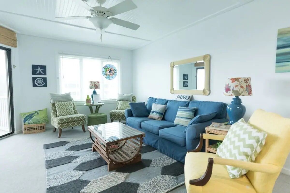 A living room with a blue sofa, a yellow armchair, a glass coffee table, and a ceiling fan.