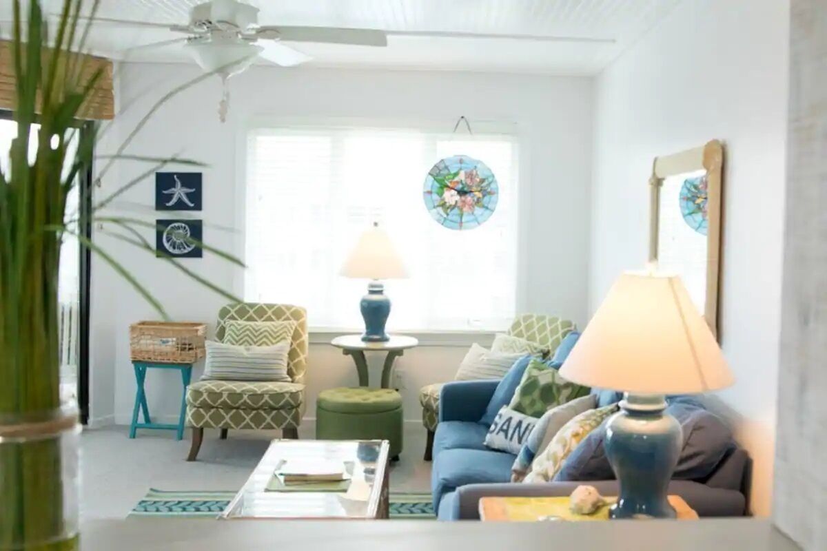 A living room with a ceiling fan, a sofa, a coffee table, and a lamp. There are decorative items on the walls and a window with a view.