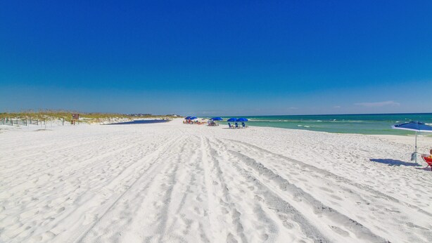 A sandy beach with footprints leading to the water, a few umbrellas, and a clear blue sky.