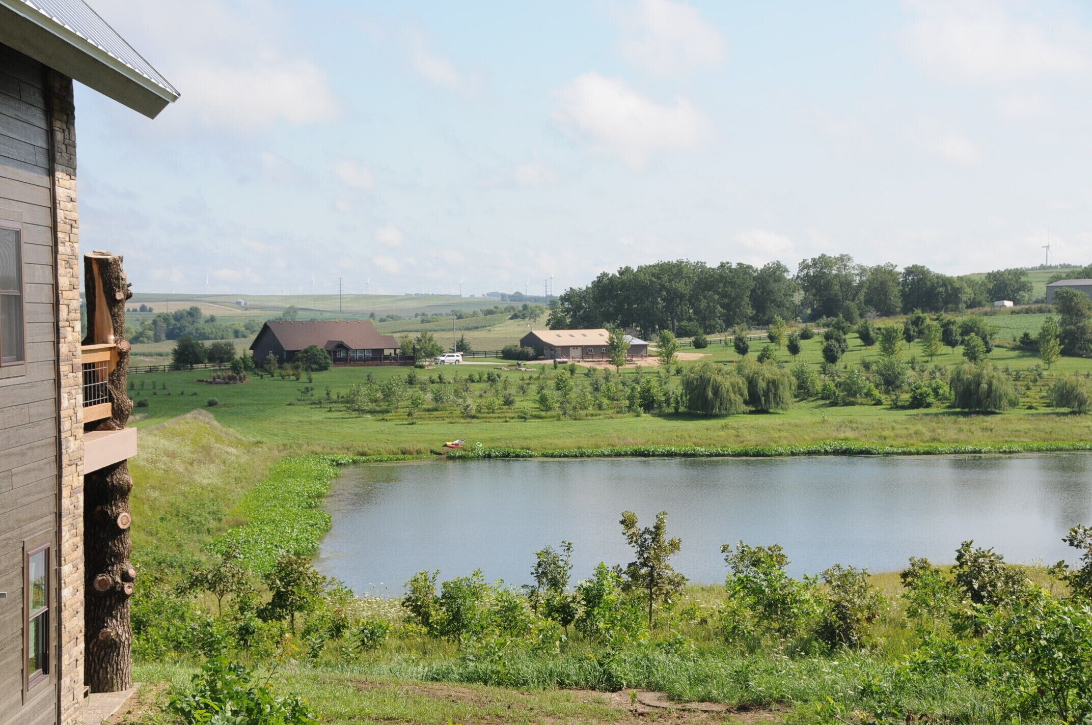A view of a house with a large pond in the foreground, surrounded by greenery and a few buildings in the background.