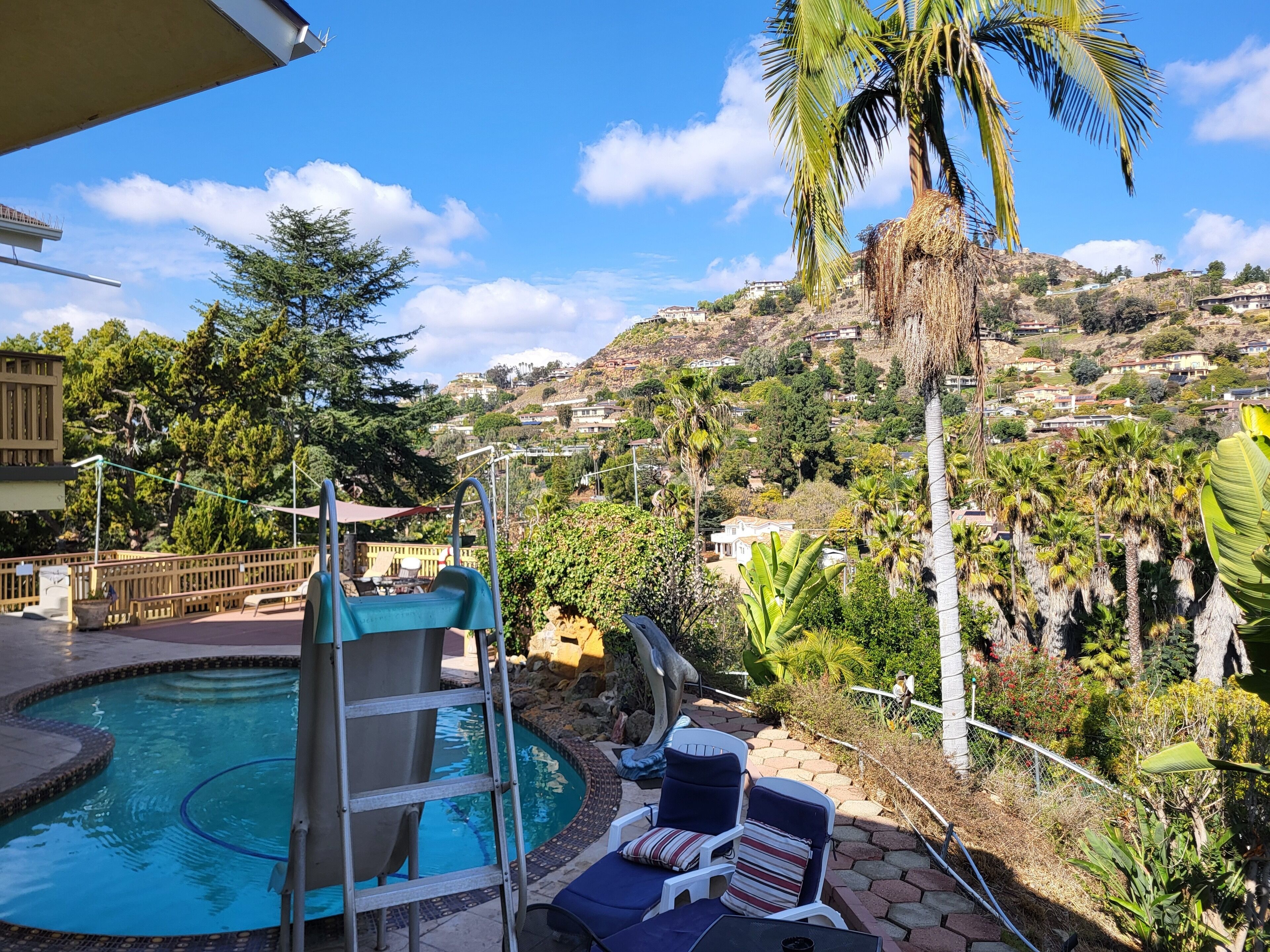 A pool area with a slide, lounge chairs, and a view of a hillside with houses.