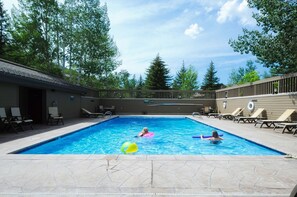 A rectangular swimming pool with two people in the water, surrounded by lounge chairs and a wooden deck.