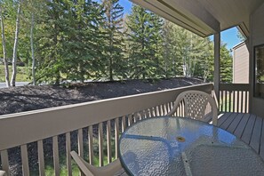 A covered outdoor deck with a glass table and chairs, surrounded by a wooden railing, overlooking a forested area.