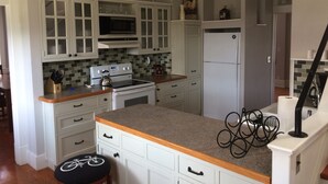 Een keuken met witte kastjes, een fornuis, een magnetron en een koelkast. Er is een houten aanrecht en een fietsdecoratie op de stoel.