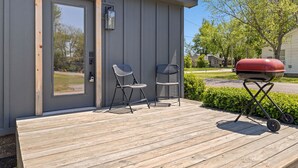 Una casa pequeña y moderna con una terraza de madera, dos sillas metálicas y una parrilla.