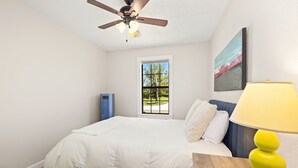 Un dormitorio con cama, ventilador de techo, ventana, un cuadro y una lámpara.