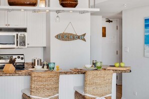 A kitchen with white cabinets, a microwave, a stove, wicker chairs, a hanging fish sign, and a door leading to another room.