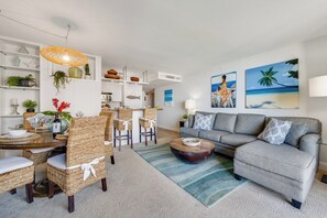 A modern living room with a grey sofa, wicker chairs, a dining table, and artwork on the walls.