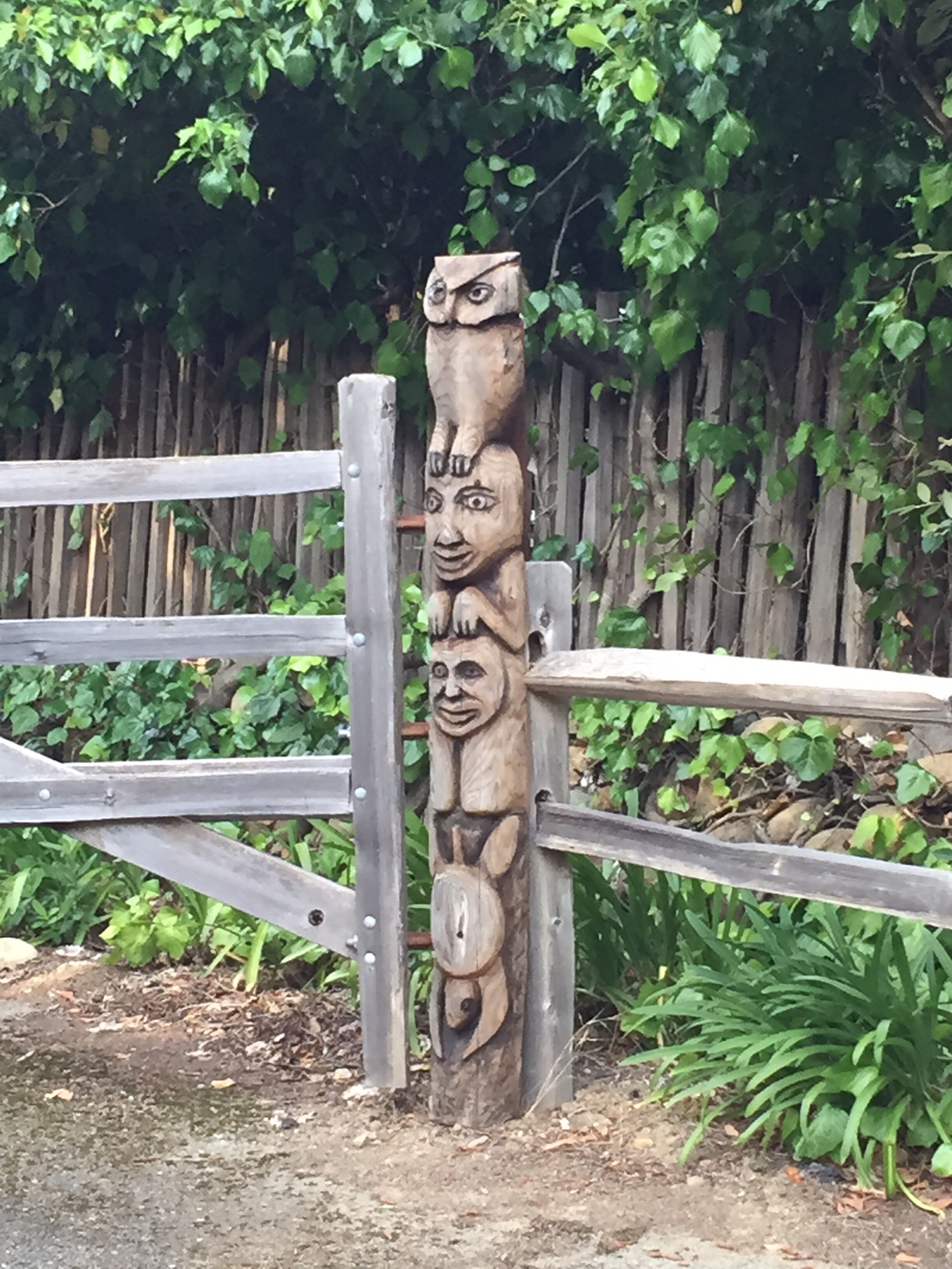 Un totem orné de trois visages, une clôture en bois et de la verdure en arrière-plan.