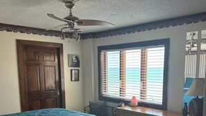 Quarto com cama, escrivaninha, ventoinha no teto, janela com persianas e um cesto de lixo.