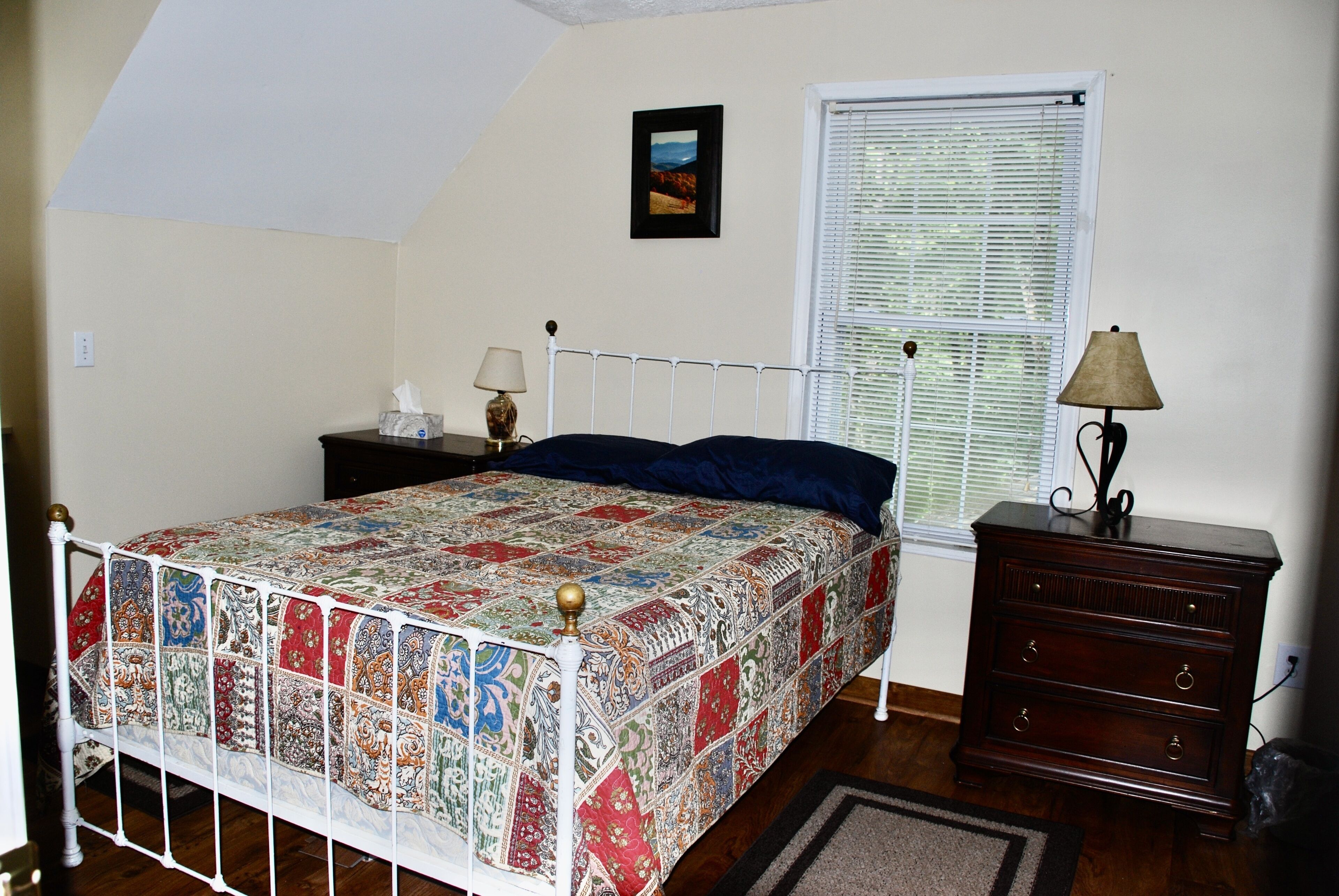 A bedroom with a bed, a dresser, a nightstand, a lamp, and a window with blinds.