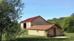 Un granero de madera con techo rojo y una estructura más pequeña adosada, rodeado por un campo cubierto de césped y árboles.