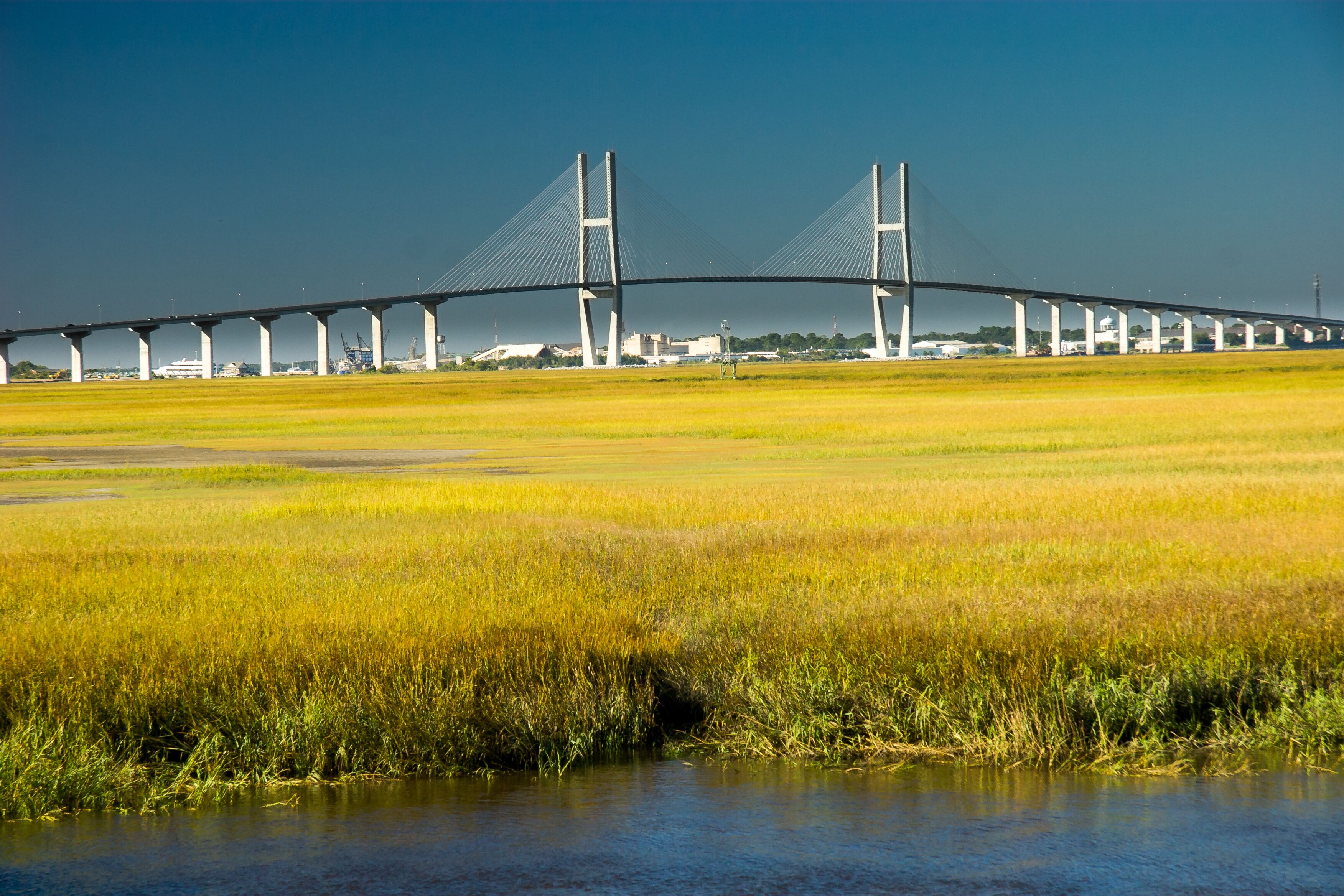 A large suspension bridge spanning a wide river with a flat landscape in the foreground.