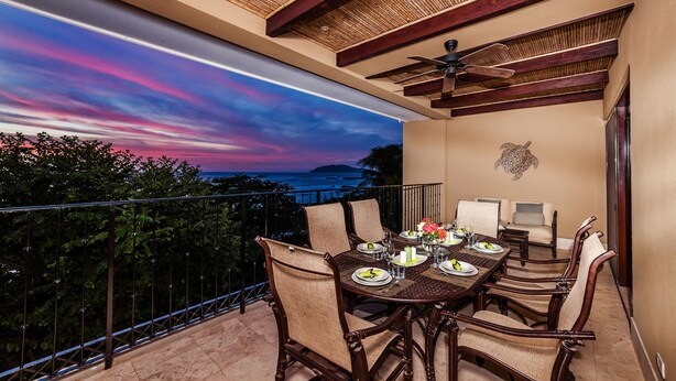 A dining area with a table set for four, surrounded by chairs, overlooking a scenic view of the ocean and hills.