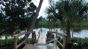 Una terraza de madera con una silla y una mesa blancos, con vista a una masa de agua tranquila con árboles y una palmera al fondo.