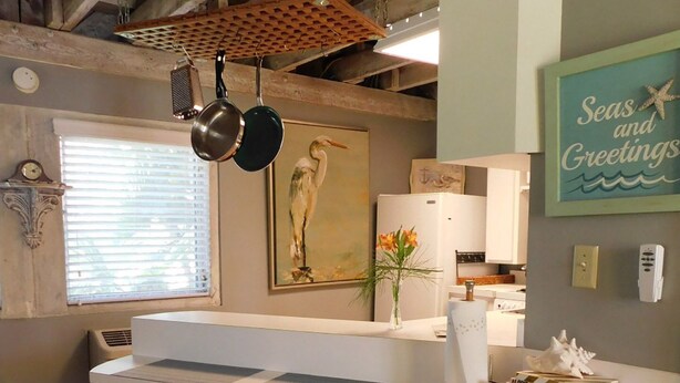 Una cocina con techo de madera, encimera blanca, dos taburetes de madera, un florero con flores y un cuadro con la imagen de un pájaro.