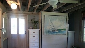 Una habitación con techo de madera, un aparador blanco con cajones, un cuadro de un velero y una puerta con paneles de vidrio esmerilado.