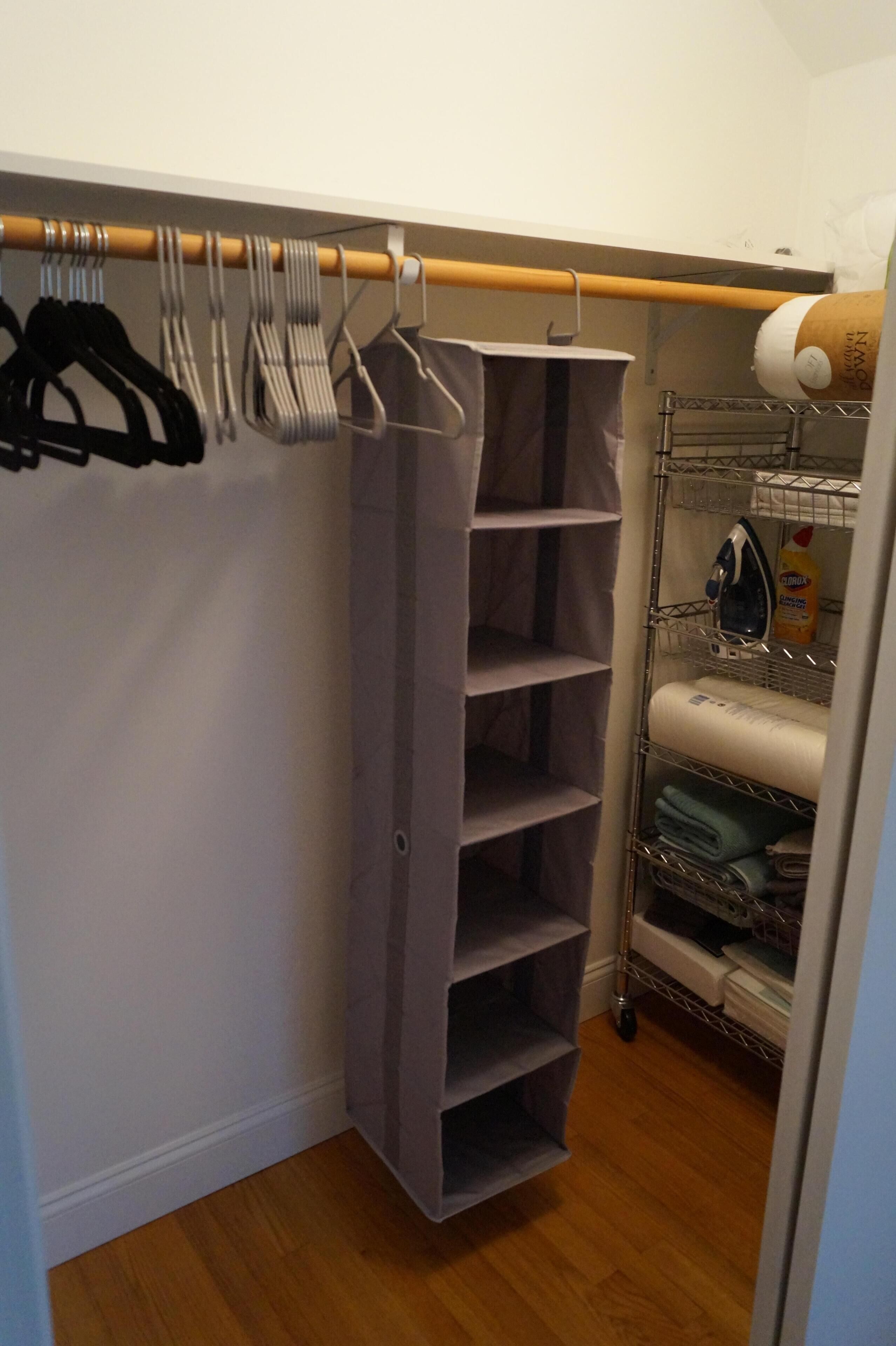 A closet with hanging clothes, shelves, and a storage unit with folded clothes and cleaning supplies.