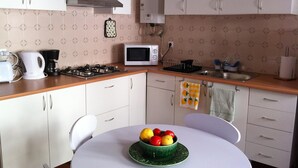 A compact kitchen with white cabinets, a microwave, a stove, and a sink. There's a table with a bowl of fruit and a kettle. - Country and  beach, the best of both worlds. (Famalicão da Nazaré)