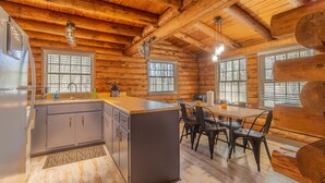 L’intérieur d’une cabane en rondins comprend une cuisine et un coin repas, avec des poutres en bois, un îlot central et une grande fenêtre.