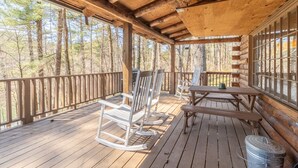 Une terrasse en bois, agrémentée de fauteuils à bascule et de tables de pique-nique, entourée d’un chalet en bois doté de grandes fenêtres.