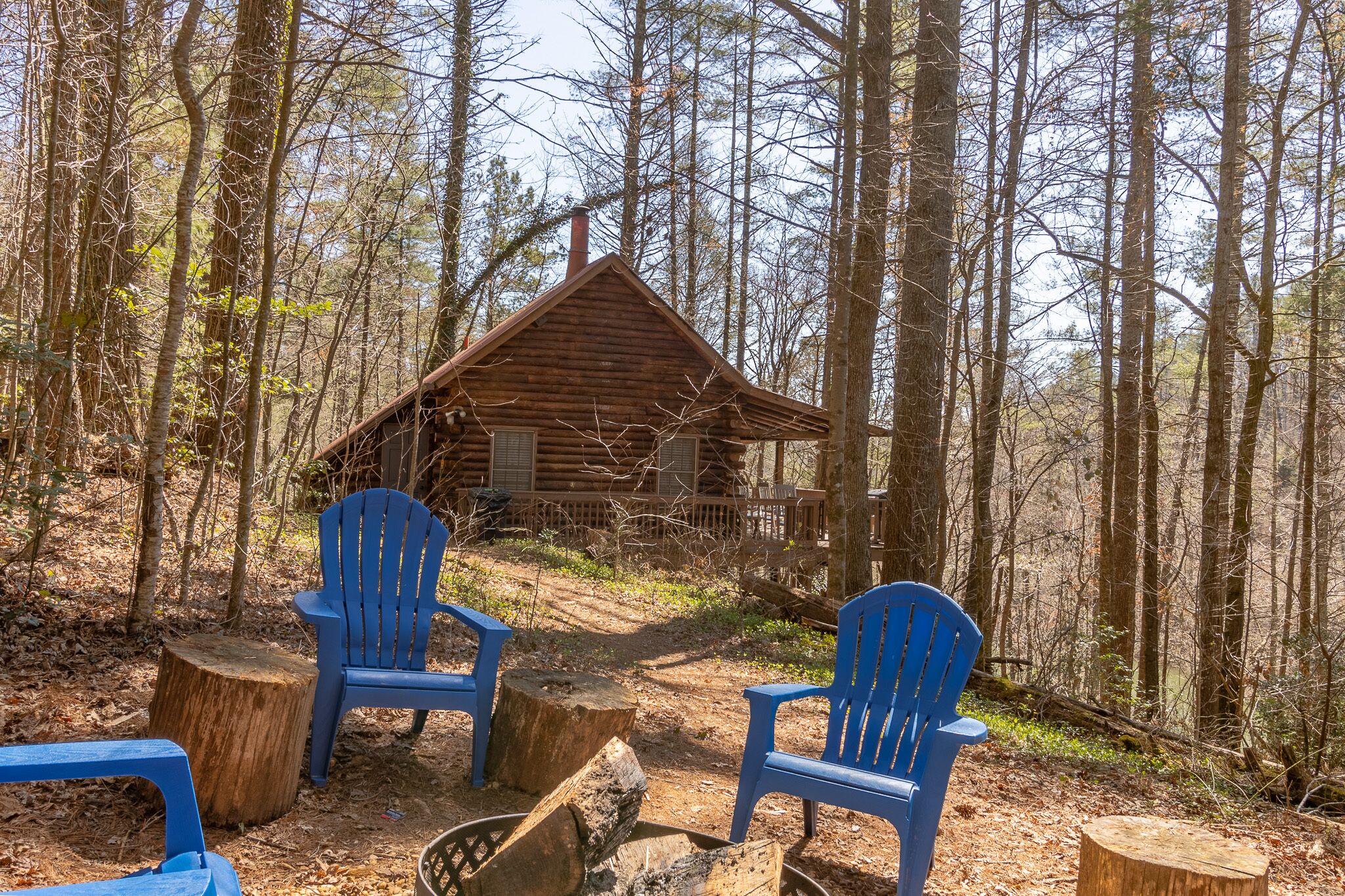 A rustic cabin surrounded by tall trees, with two blue chairs and a fire pit in the foreground.