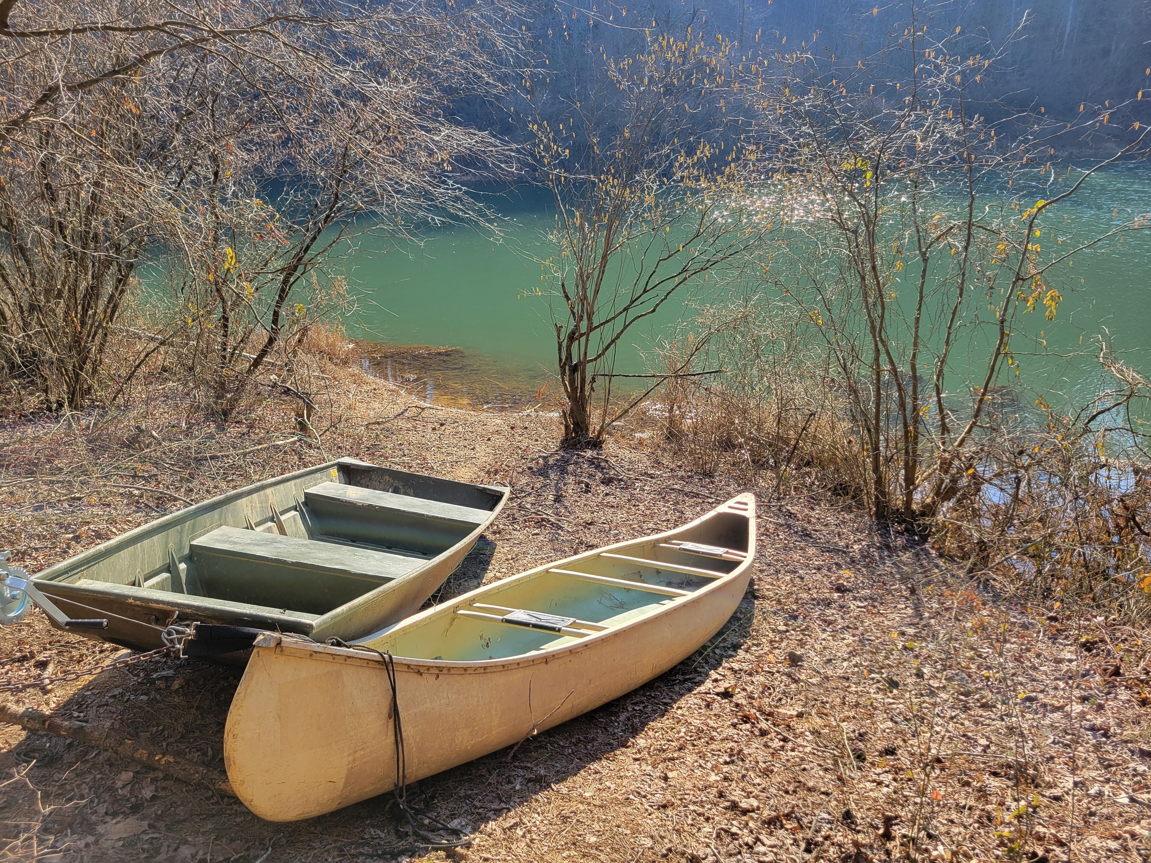 Deux canoës reposent sur un sol sec, recouvert de feuilles, près d’une étendue d’eau.