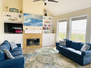 A living room with a fireplace, two sofas, a TV, and a large window with a view of the beach.