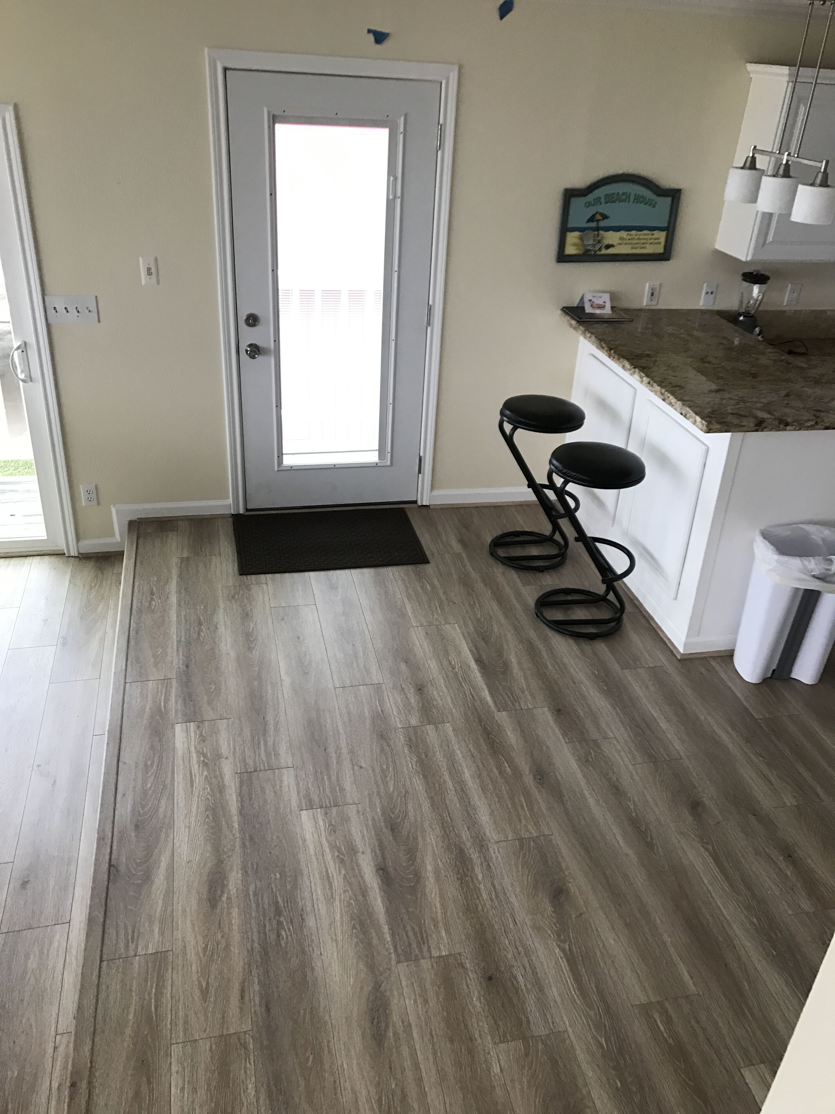 A kitchen with a door, a black doormat, a wooden floor, a bar stool, and a countertop.