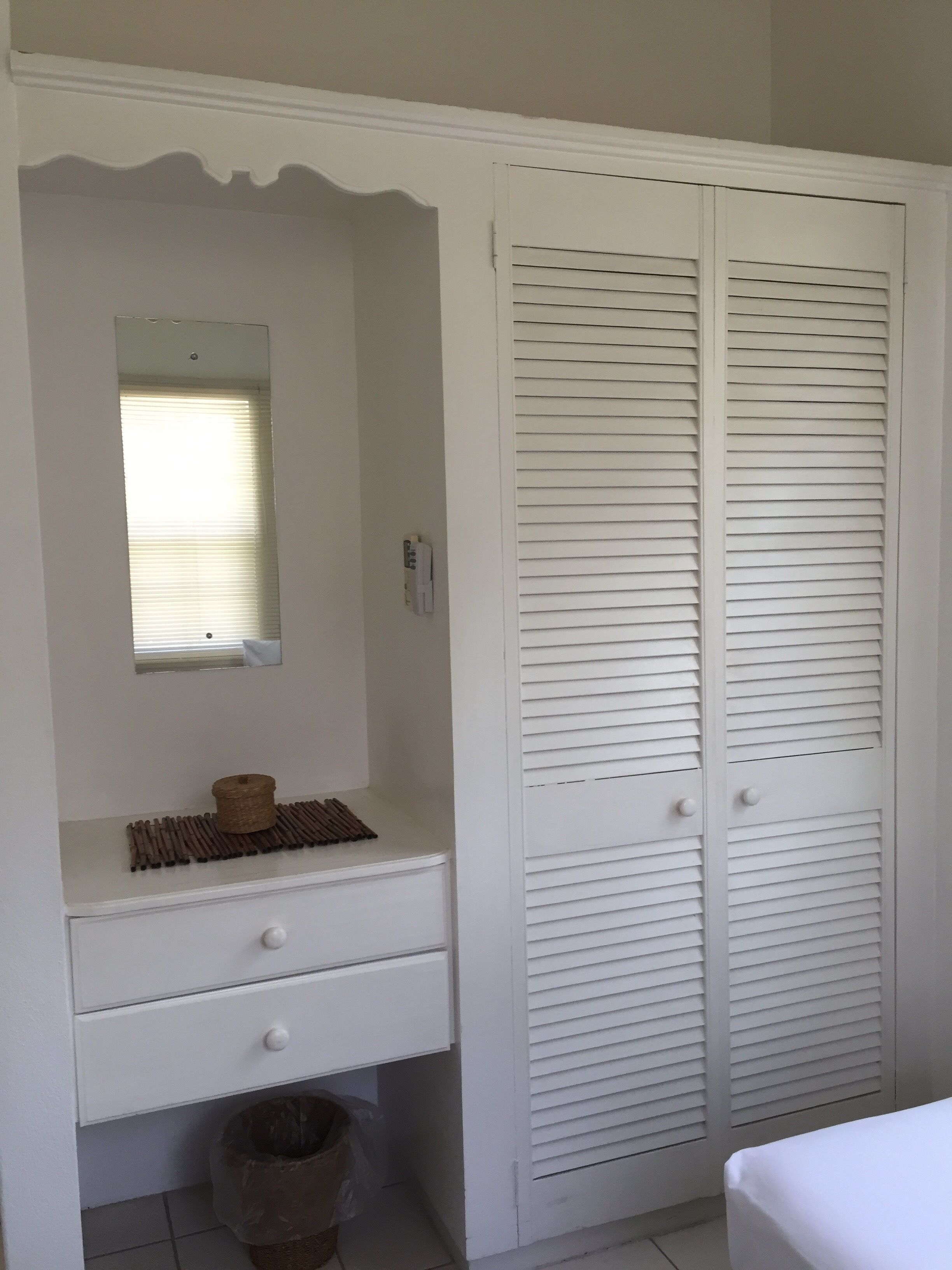 A white wardrobe with a mirror, a small shelf with a basket, and a trash bin.
