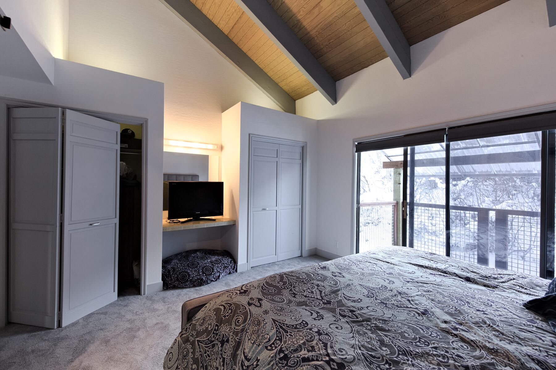 A bedroom with a large bed, a TV on a shelf, a walk-in closet, and a balcony with a view.