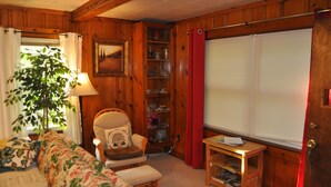 Een knus interieur met een houten lambrisering, een comfortabele bank, een stoel, een tafeltje met een lamp, en een raam met jaloezieën.