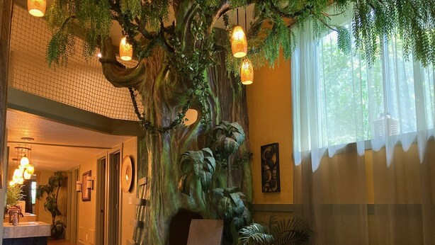 Un dormitorio con un mural grande de un árbol, una cama, una silla y un escritorio con una lámpara.