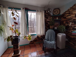 Une pièce avec un sol carrelé, une chaise recouverte d’une couverture, un radiateur, un mur en bois orné d’une horloge et une fenêtre avec des rideaux.