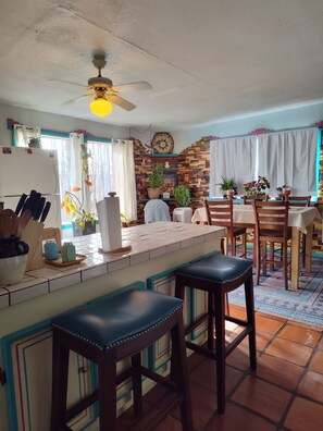 Una cocina con una isla central, taburetes de bar, un ventilador de techo y una zona de comedor con sillas y mesa.