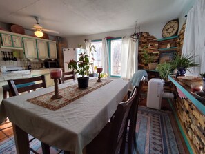 A dining area with a table set for two, chairs, a ceiling fan, a kitchen area with cabinets, a refrigerator, and a window with curtains. - Casita Rael, an Art lovers get-a-way (Taos)