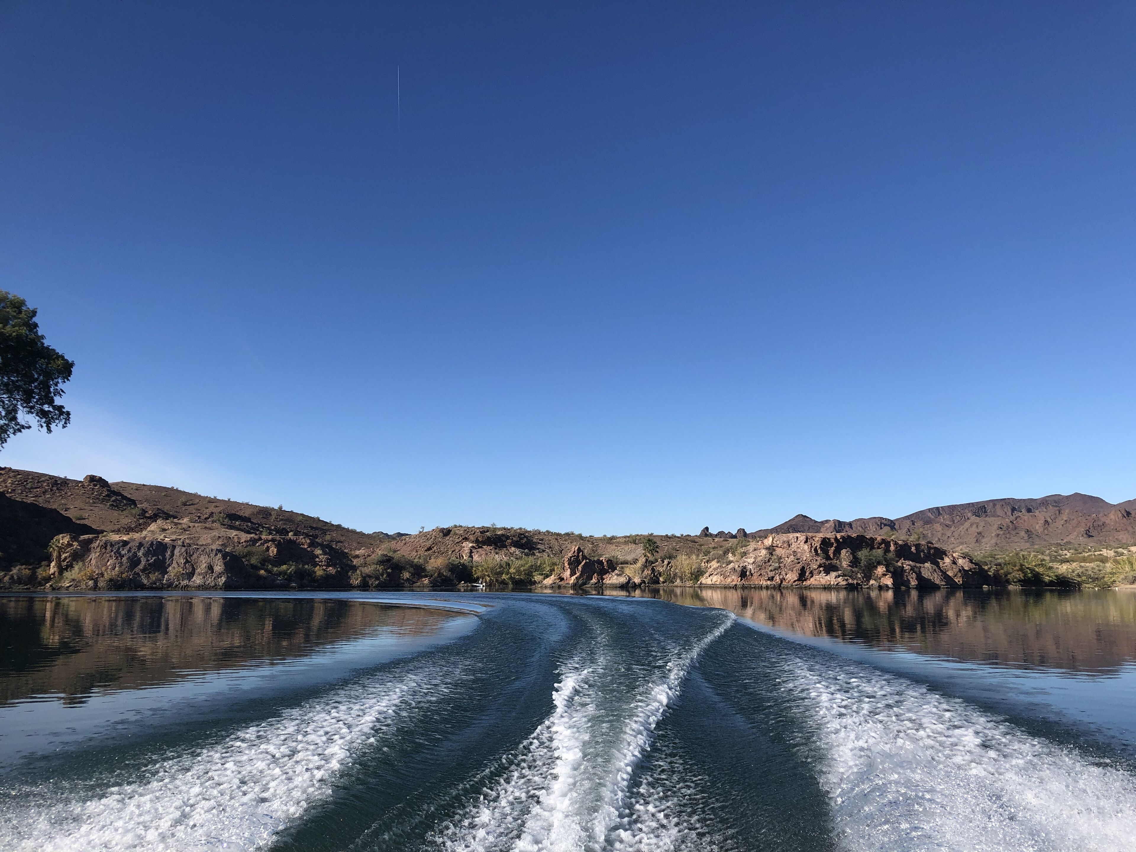 Vista despejada de un lago con estela tras un barco en movimiento, rodeado de colinas rocosas y vegetación escasa.