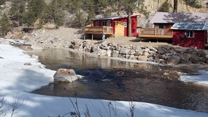 Une cabane dotée d’une terrasse en bois et d’une façade rouge, située au bord d’une rivière dont les rives sont rocheuses.