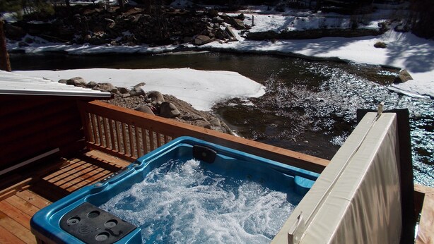 Un bain à remous sur une terrasse en bois, avec vue sur une rivière et des rochers enneigés.