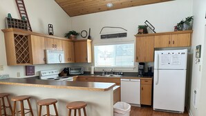 Une cuisine moderne avec des armoires en bois, une cuisinière encastrée, un lave-vaisselle et un réfrigérateur. On y trouve également des tabourets en bois et une poubelle.