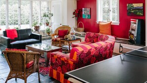 Un salon avec une table de ping-pong, un canapé noir, un canapé rouge, des fauteuils en osier et un mur rouge.